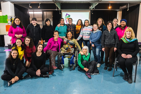 The entire group of workshop participants pose for the camera in an enclosed room at the Fontanarrosaa Cultural Center, with a plain blue floor. In the background, the screen showing the logo of Desear con Inclusión (a map of Argentina in green, orange and purple). In the second line, twelve women are smiling, standing, facing the camera. Some are blind, others are deaf, others have multiple disabilities or psychosocial disabilities. There are also the two Radio Frida operators.