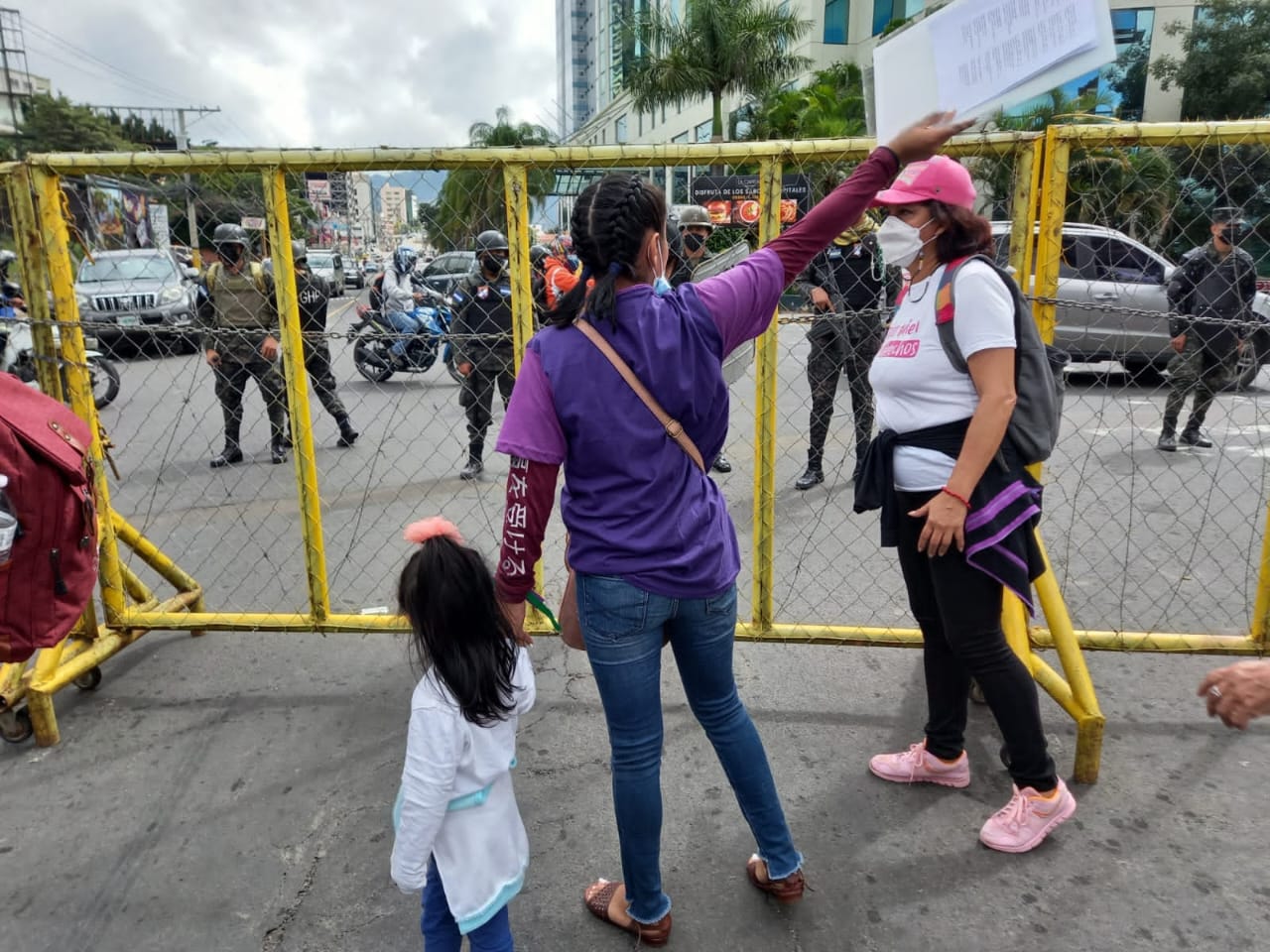 One woman holding a child's hand and carrying a sign in her other hand is talking to another woman wearing a mask. Military officers can be seen in the back, behind a fence.