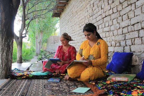 Two women seating on a rug outside writing in notebooks