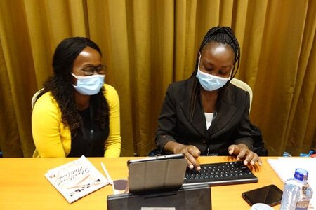 Two black women wearing masks sitting next to each other and looking at a tablet on a table