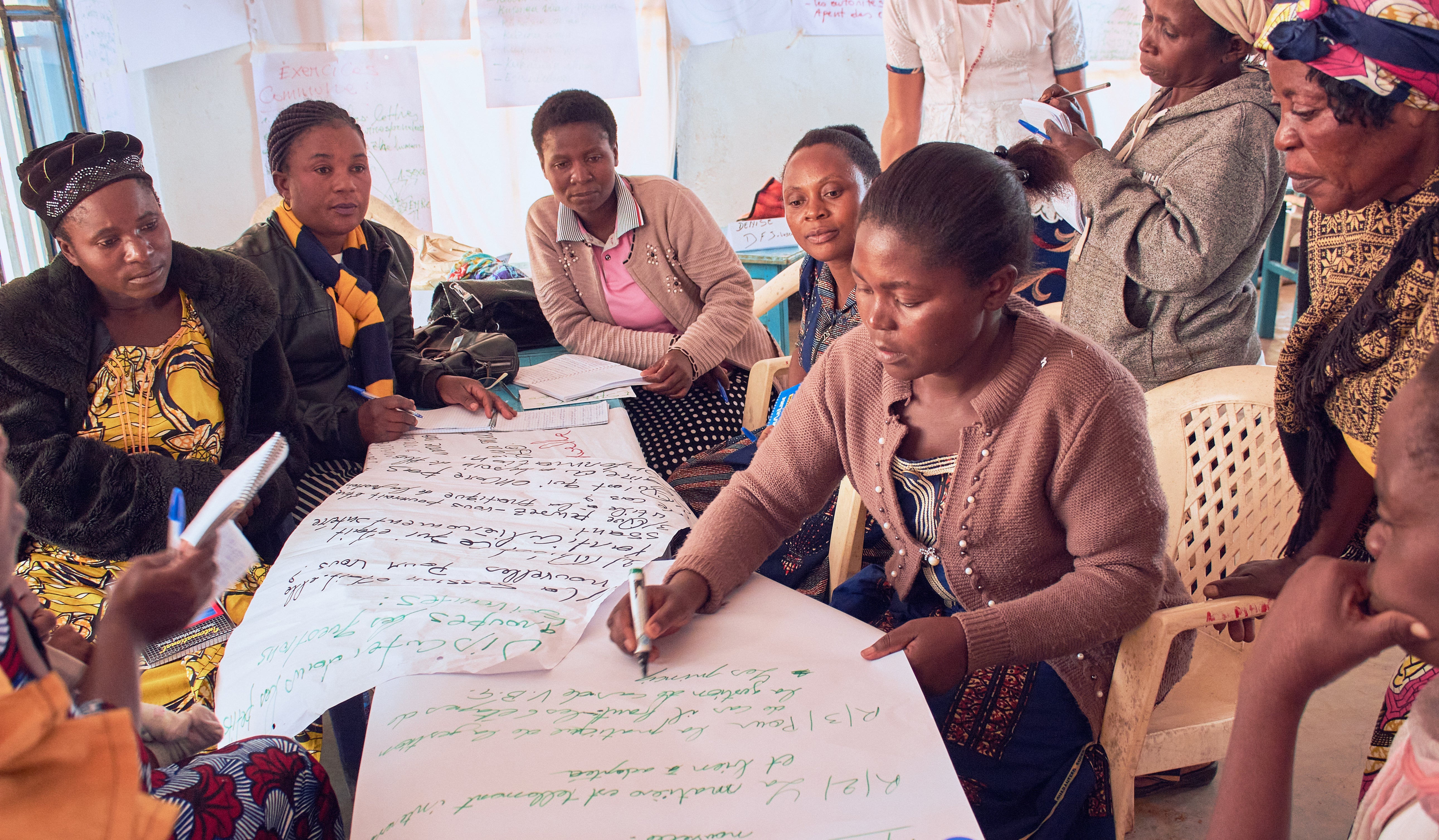 A group of black women is seen sitting at a table, they are holding and writing on two large pieces of white paper