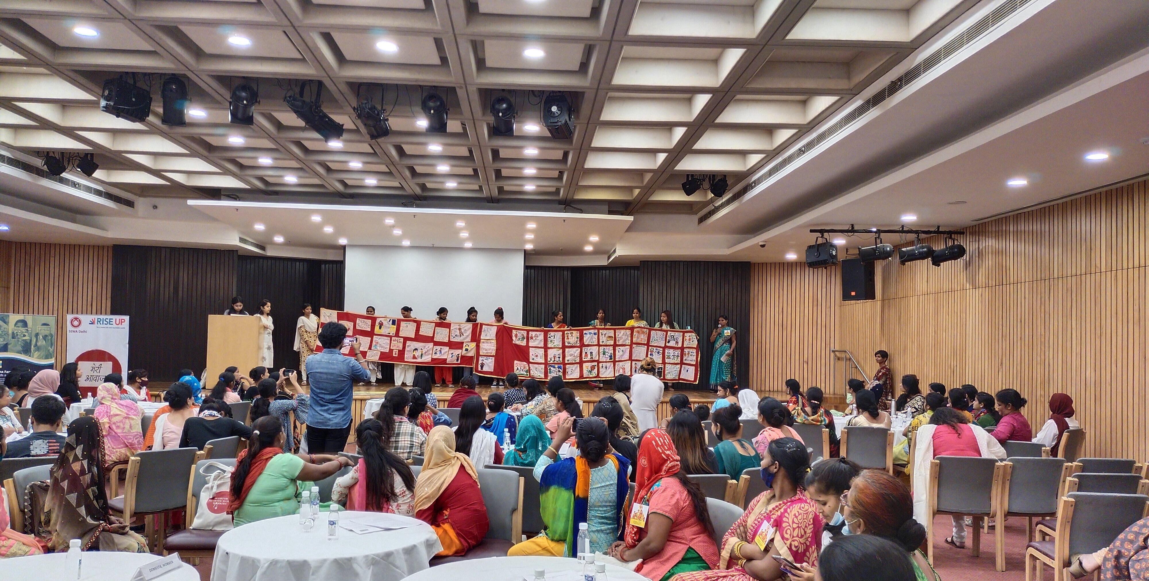 Large group of women domestic workers in a conference room looking at women on a stage showing a very long piece of red tissue (sari) where they stitched their experiences of sexual harassment in the workplace.
