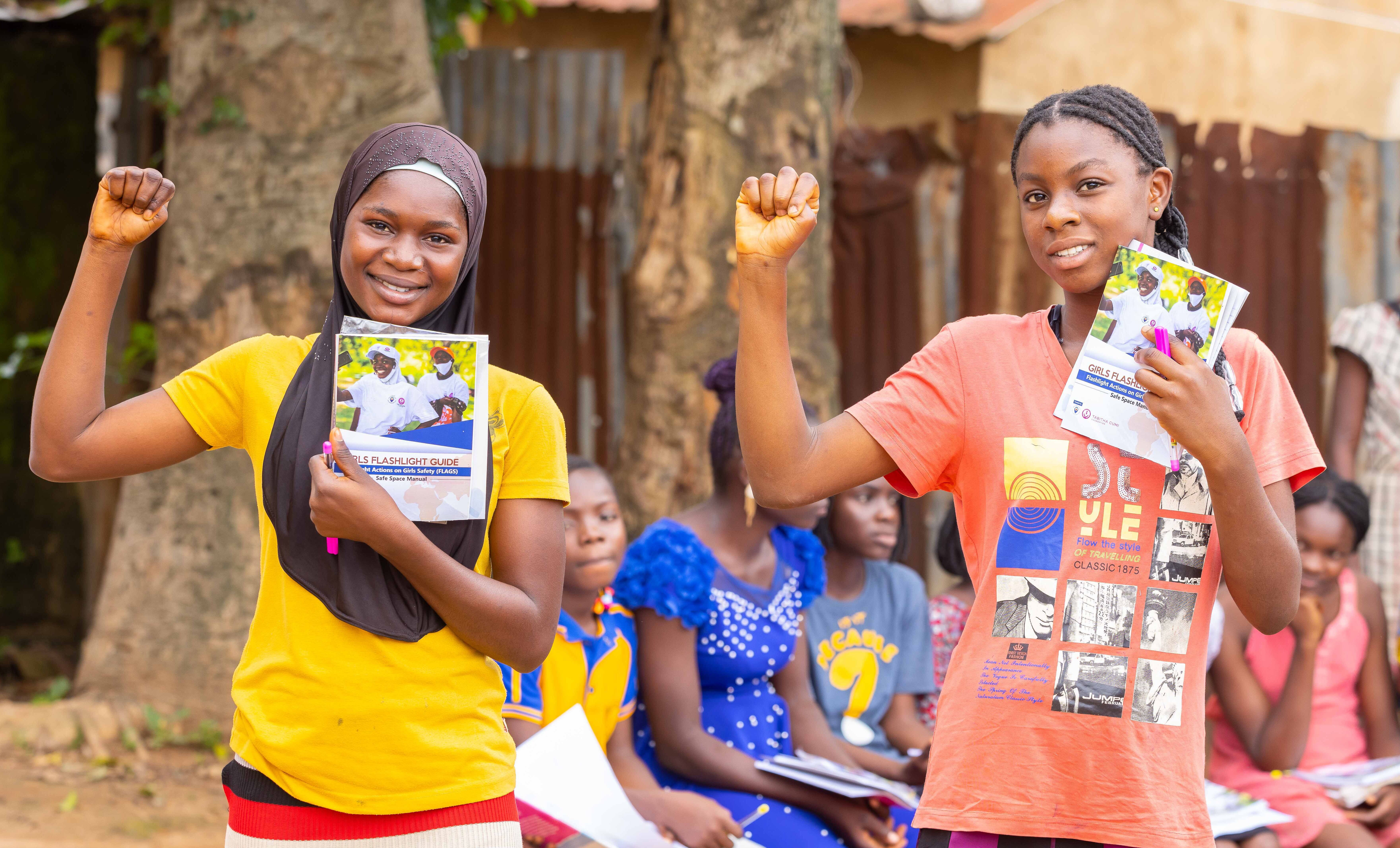 Two young black girls are raising their fist while holding up a flyer with the other hand. One is wearing a yellow t-shirt, the other one is wearing an orange t-shirt. They are smiling at the camera and standing outside.