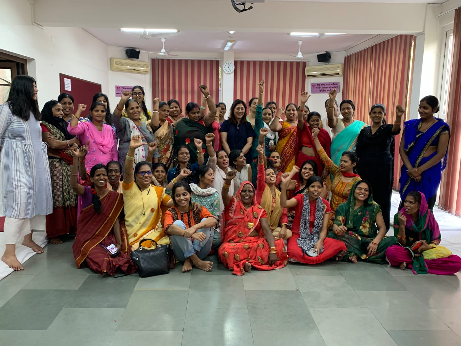 Group of women wearing colourful clothes sitting and standing in a room, facing the camera and smiling, some of them have their fists raised up in the air.