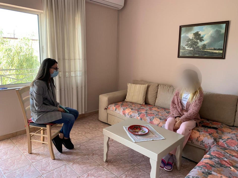 In a room, one woman wearing is mask is sitting on a chair facing another woman with a blurred face, sitting on a large collared couch.