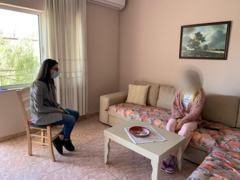 Two women are seen talking. They are in a room with a window on the left, one woman wearing a mask is sitting on a chair facing the other woman, sitting on a large couch. Her face is blurred to protect her identity.