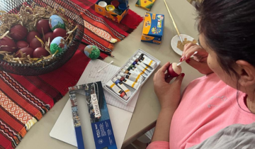 A woman wearing a pink t-shirt, with brown hair, is seen painting on an egg above a table. Her face is not visible.