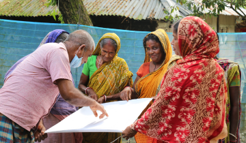 A group of people, both men and women, are holding a large piece of white paper. One man is pointing at the paper, a woman is seen smiling. They all are wearing colourful clothes, and are standing outside.