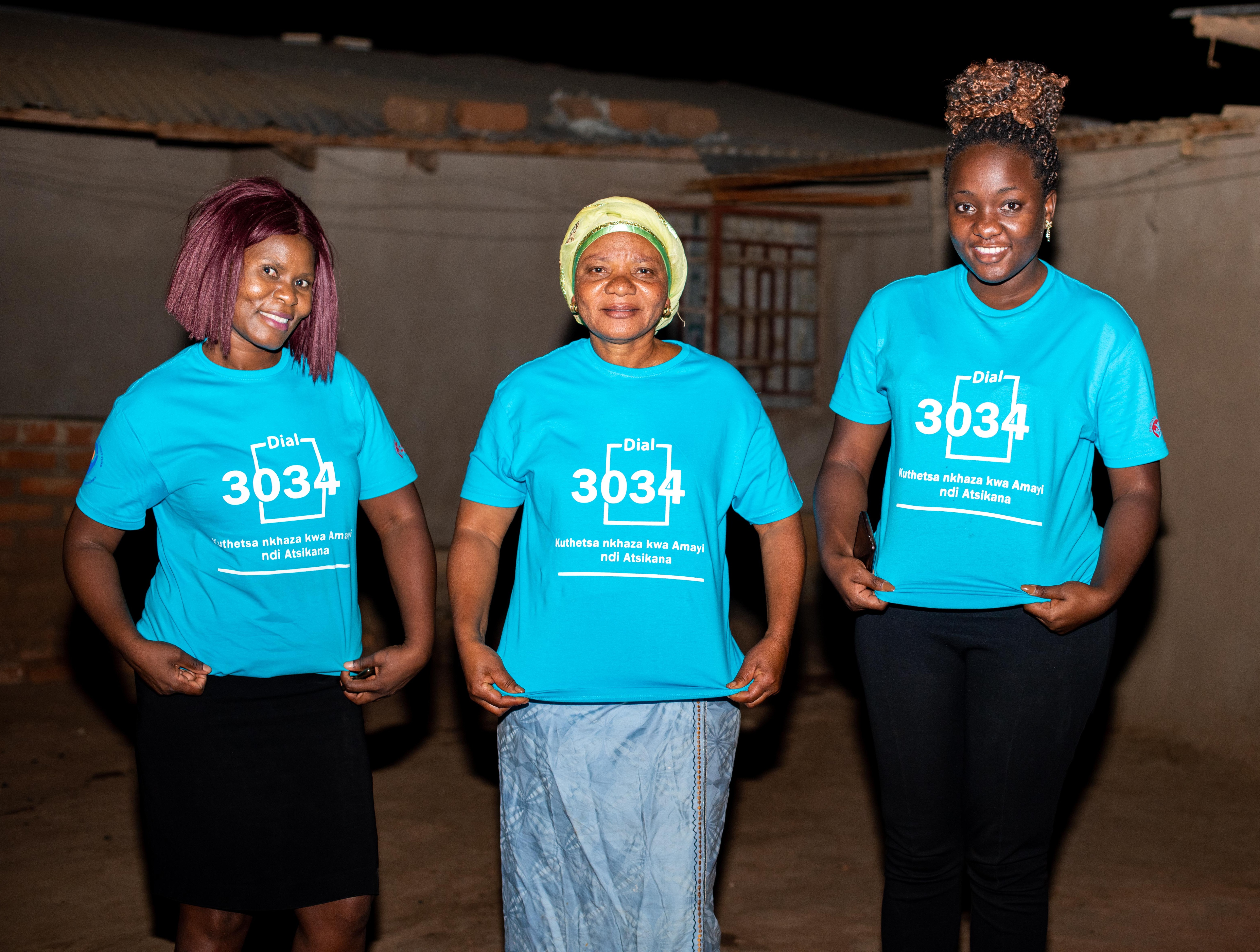 Three black women outside in the dark, of all ages, are showing their blue t-shirts with the number of FACT Malawi's new hotline 