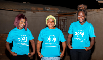 Group of three black women outside at nighttime, showing off their blue t-shirts, all identical, with the hotline number of FACT Malawi