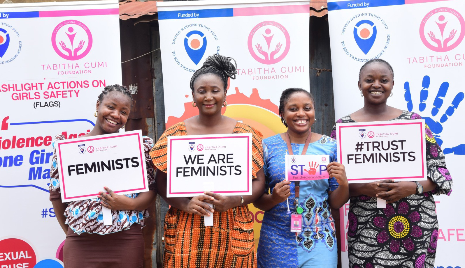 Four black women are standing outside, smiling, holding each a poster. One says "FEMINISTS", another one says "WE ARE FEMINISTS", then a smaller poster reads "STOP violence", and the last poster is the hashtag TrustFeminists