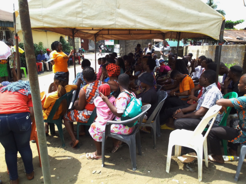 Group of people sitting outside underneath a tent, listening to a woman standing in front of them and speaking to them