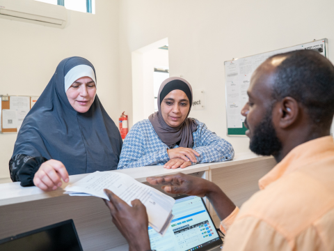 Two women are talking to a man standing behind a desk, one of the woman is handing him out a piece of paper.