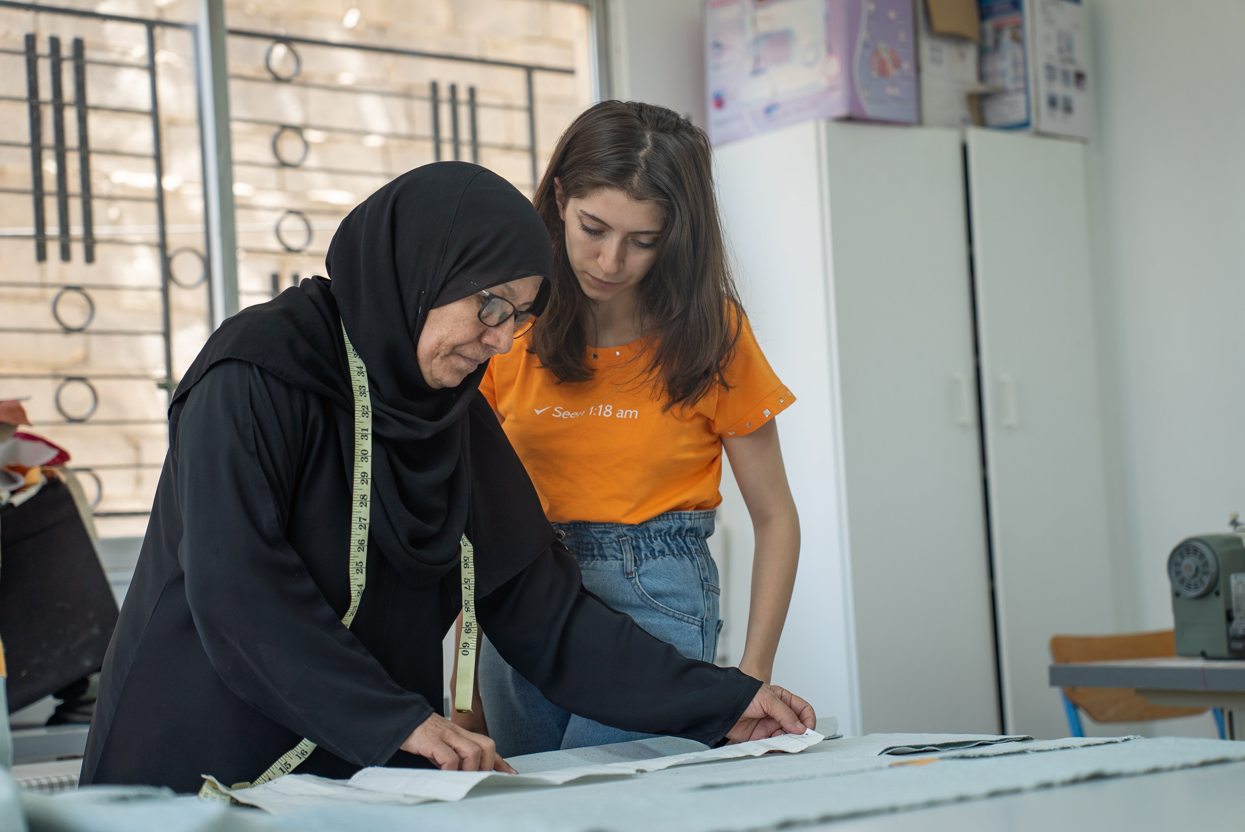 One older woman is showing a piece of a paper to a young woman who is wearing jeans and an orange t-shirt.