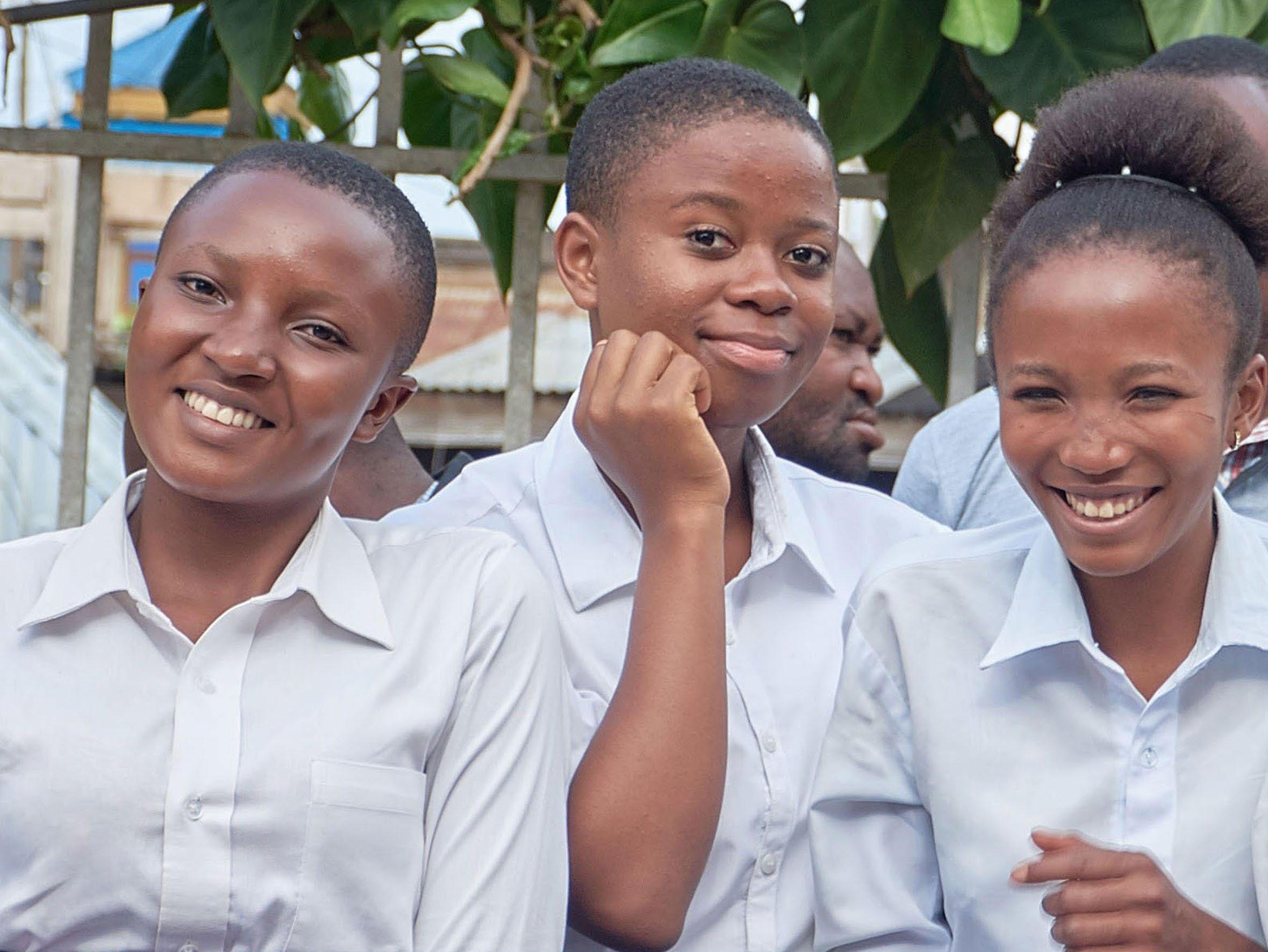 Three young black girls are wearing school uniforms and smiling at the camera