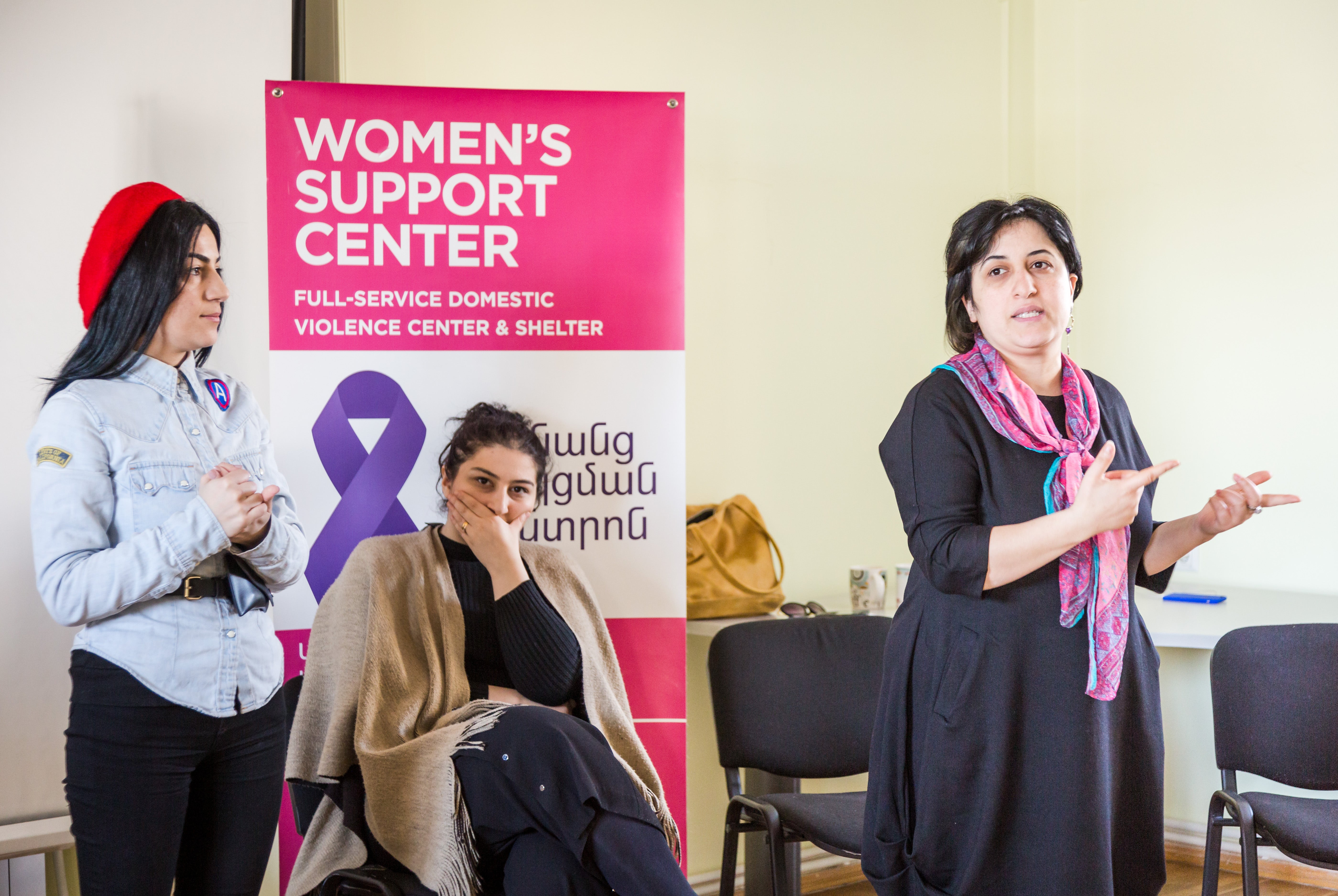 Two women are standing in a room, one is speaking. Another woman is sitting on a chair, holding her hand over her mouth, in front of a large banner that reads Women's Support Center in white on a pink background