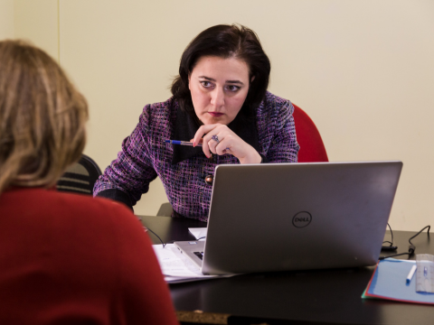 A brunette woman sitting at a desk in front of a computer is looking up at another woman, seen from the back, sitting in front of her on the other side of the desk