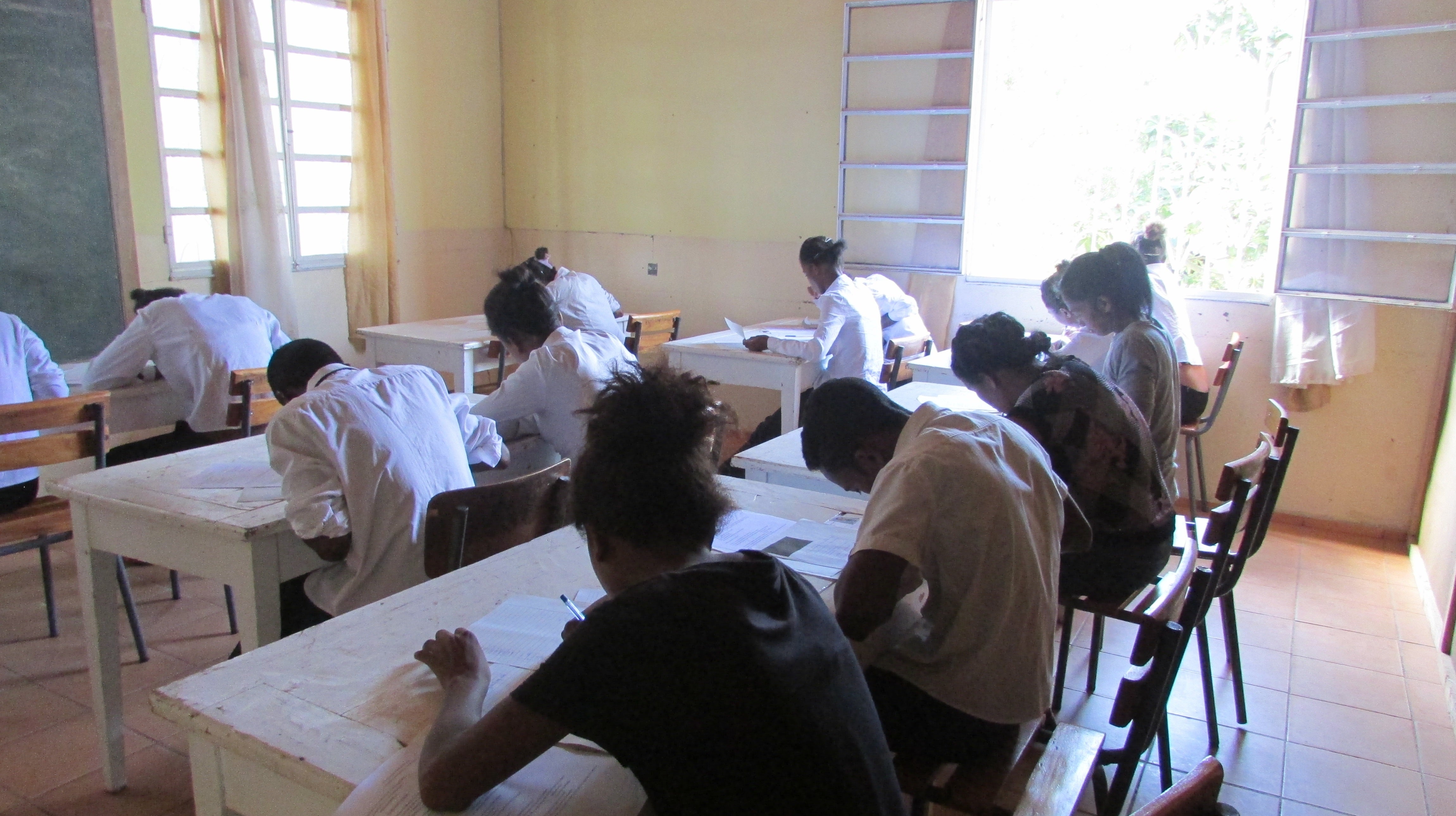 Group of young people sitting at tables and writing, their back is to the camera, they seem to be inside a classroom
