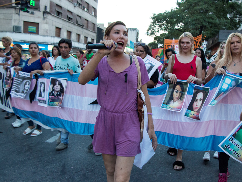 photo shows a group of people marching with one person holding the microphone and the people behind holding a large banner in the colors of transgender flag (pastel blue and pastel pink)