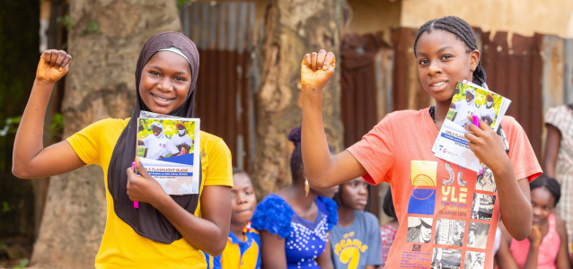 Two young girls are seeing carrying a flyer in one hand and raising their fist in the other hand. One is wearing a bright yellow t-shirt, while the other on the right is wearing a orange/red t-shirt.